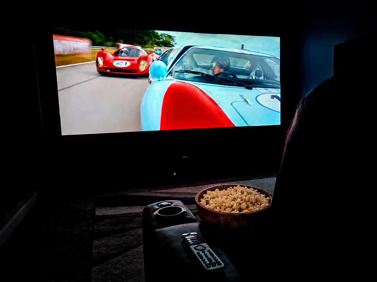 Dark home theater room with Ford v Ferrari on screen, theater seats and remote in the foreground